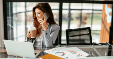 Uma mulher está no ambiente de trabalho. Ela usa o celular, sorri e olha pela a janela. Na sua mesa, há um "notebook", papéis e uma agenda. Ela segura um copo de café na mão.