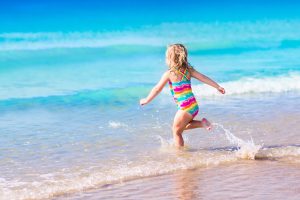 Happy child running and jumping in the waves during summer vacation on exotic tropical beach. Holiday on ocean coast for family with young children. Kids play at the sea. Little girl learning to swim.