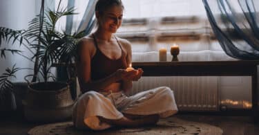 Mulher sorridente se preparando para meditar. Ela segura uma vela acessa e está rodeada por outras velas dispostas no chão em que está sentada.