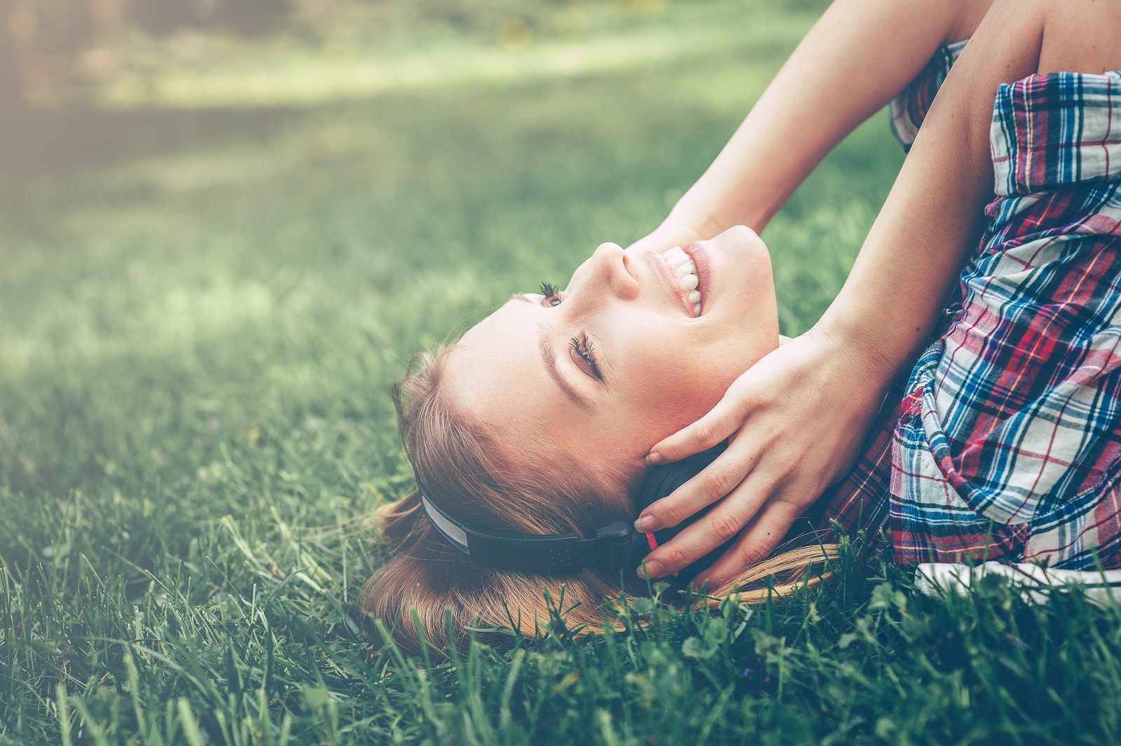 Enjoying her favorite music. Side view of beautiful young woman in headphones listening to the music and smiling while lying on the green grass