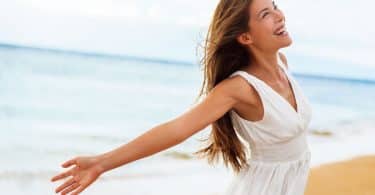 Mulher de vestido na praia, sorrindo com os braços abertos.