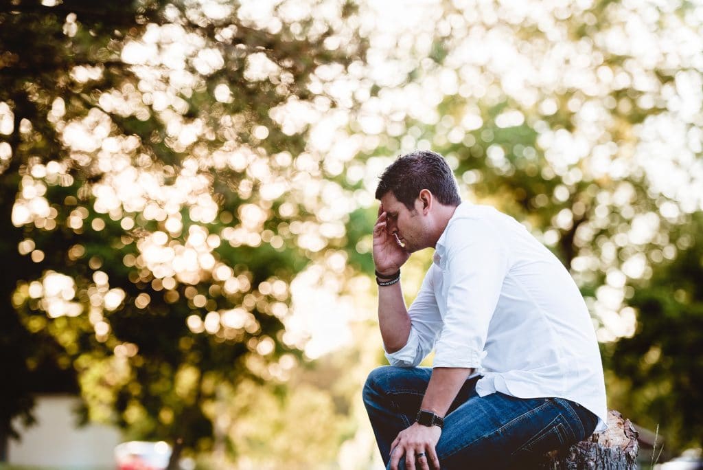 Homem sentado em um parque, com a mão sobre a testa, em sinal de cansaço.
