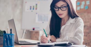 Mulher branca de óculos sentada em frente à uma mesa de escritório escrevendo num caderno.