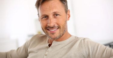 Homem com alguns cabelos grisalhos, barba rala e sorridente, sentado em sofá.