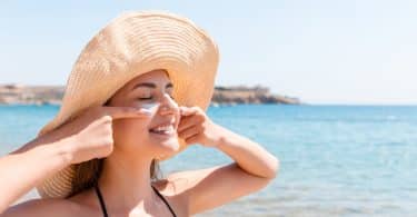 Mulher passa protetor solar no rosto. Ela está de olhos fechados, sorri e veste um chapéu. O ambiente é de praia.