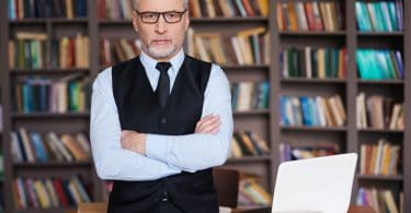 Professor em pé em biblioteca. Ele se apoia em uma mesa cheia de livros. Usa óculos e tem cabelo grisalho.