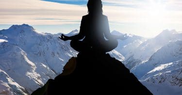 Silhueta de uma mulher sentada no pico de uma montanha meditando.