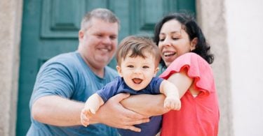 Família composta por mãe, pai e filho. Os pais seguram o filho como se estivesse voando. A mãe é morena e usa uma blusa vermelha. O pai é grisalho e usa uma blusa azul. O filho é moreno e tem o cabelo de tigela e também usa uma blusa azul.