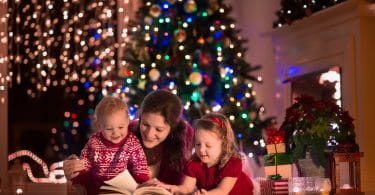 Mãe com dois filhos pequenos sentados no meio da sala com decorações de natal, lendo um livro com as crianças.