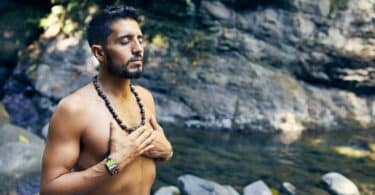 Homem meditando com as mãos sobre o peito nu. Ele está sobre as rochas que permeiam um riacho.