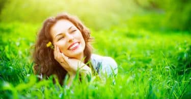 Mulher na grama com mãos o rosto sorrindo e sol refletindo