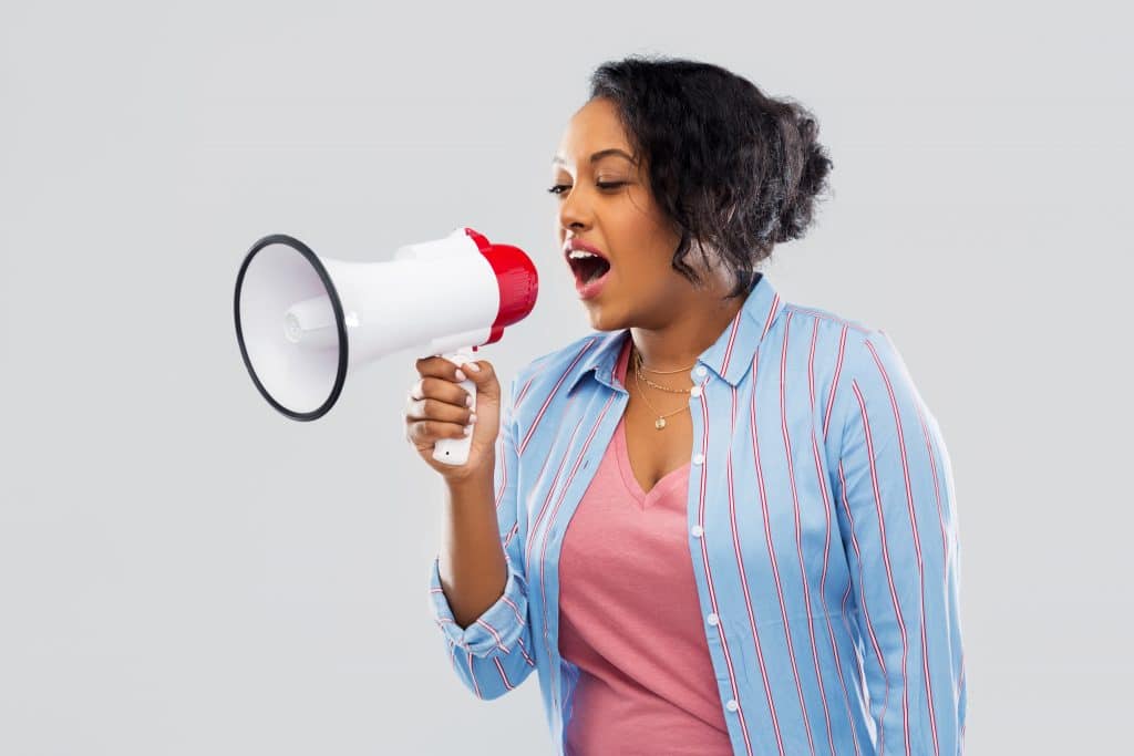 Mulher negra vestindo uma camisa azul clara com listras vermelhas segurando um megafone nas cores branco e vermelho. Ela está falando no megafone palavras de empoderamento feminino.
