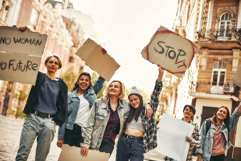 Grupo de mulheres ativistas em plena manifestação em prol do empoderamento feminino. Elas estão na rua e seguram cartazes com vários dizeres em torno do empoderamento feminino.
