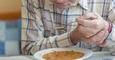 Imagem de um senhor de idade na frente de um prato de sopa levando uma colher para a boca com as duas mãos