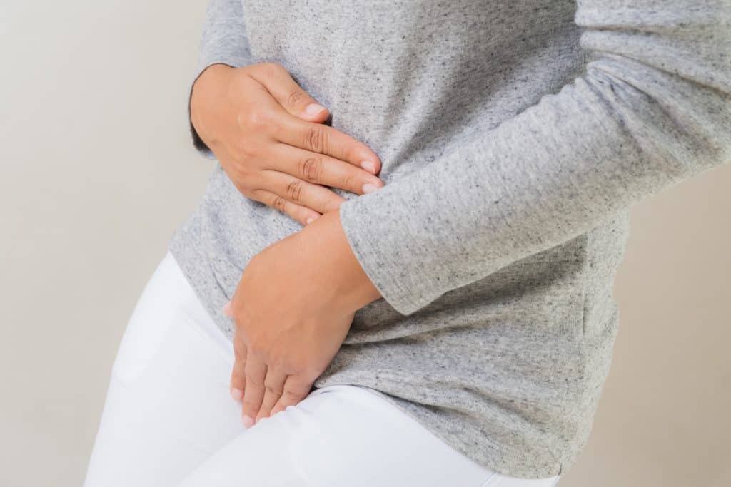 Imagem de uma mulher vestindo uma blusa de manga longa e calça branca. Ela está com as duas mãos sobre a barriga, próximo ao ventre.