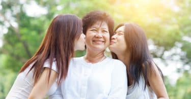 Duas mulheres beijando a mãe na bochecha, enquanto esta sorri.