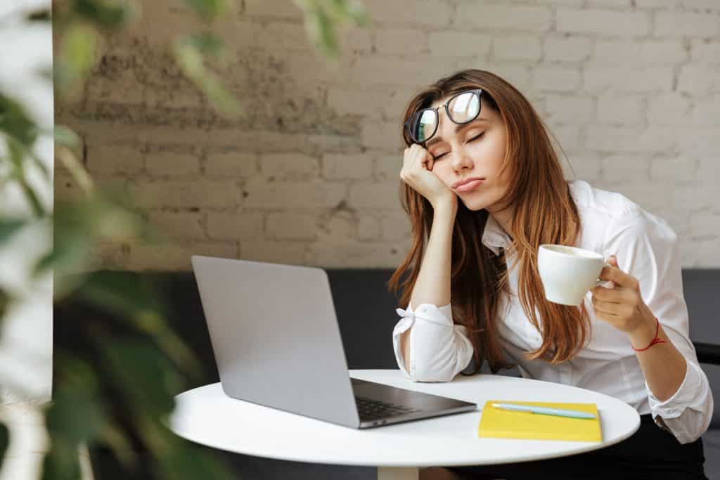Mulher em frente a um computador, apoiando seu rosto em sua mão, e segurando uma xícara de café. Seus olhos estão fechados.