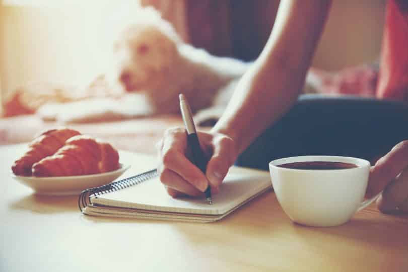 Mulher segurando uma caneta e escrevendo em seu caderno ao lado de uma xícara de café