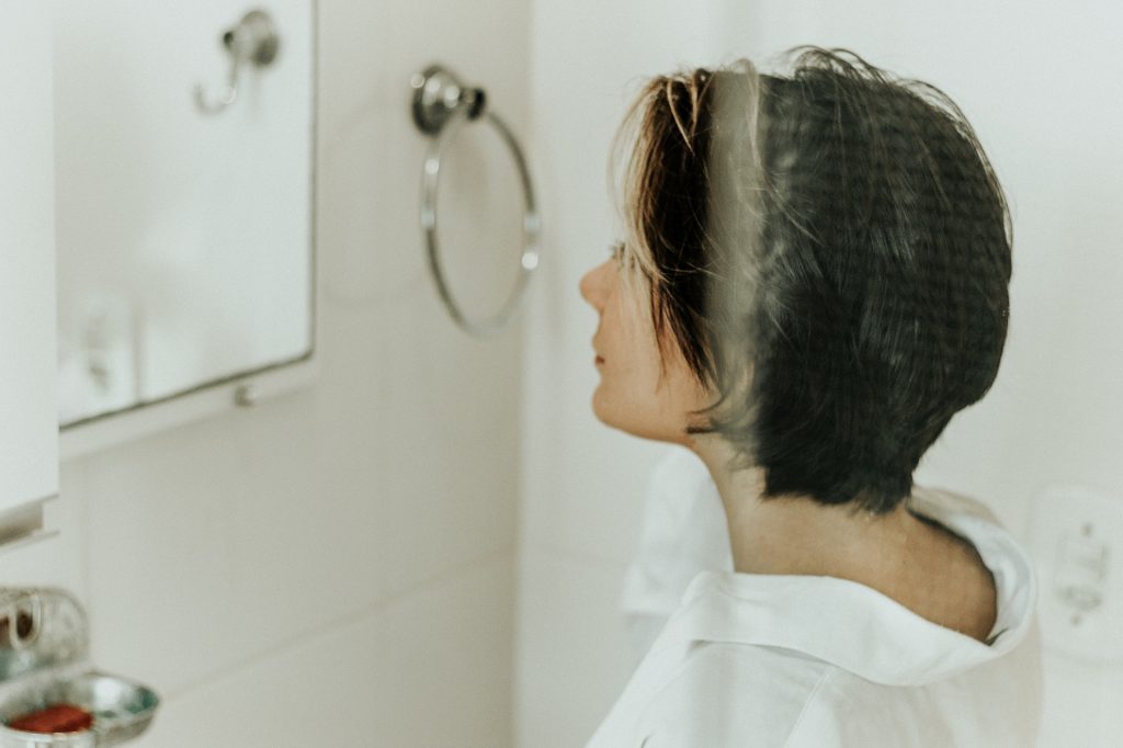 Mulher de cabelos curtos olhando para seu reflexo no espelho de um banheiro.