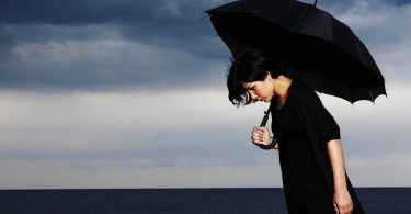 Imagem de um dia nublado na praia. Ao fundo uma mulher usando uma roupa preta e segurando um guarda chuva preto. Ela está com depressão.