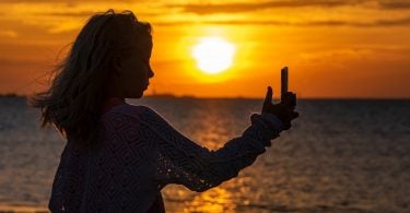 Imagem da silhueta de uma jovem fazendo uma selfie em pleno por do sol em um alto dia de verão.