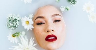 Imagem de um rosto oriental. Sobre ele algumas flores brancas, enfeitando. É uma modelo com pele branca, olhos pintados e boca com um lindo batom na cor vermelho.