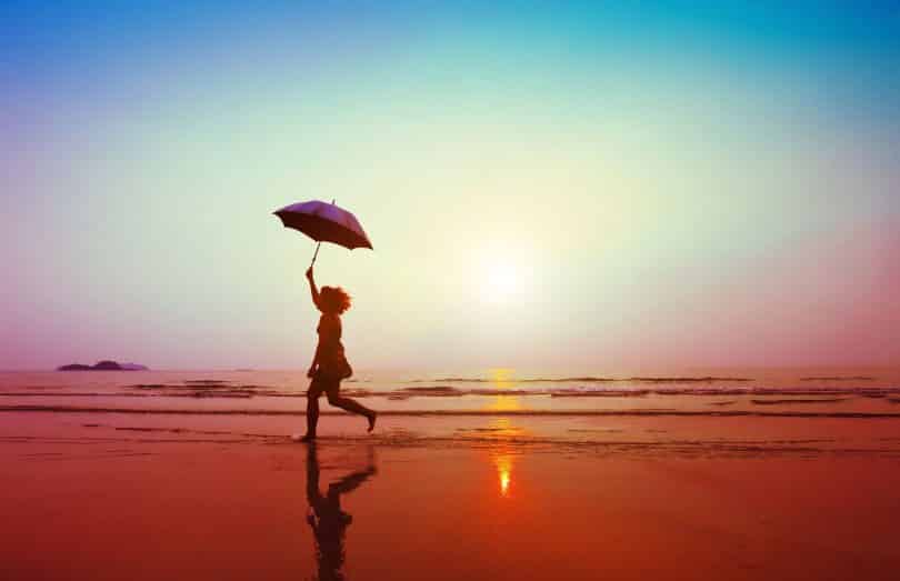 Mulher correndo na areia da praia segurando guarda-chuva.