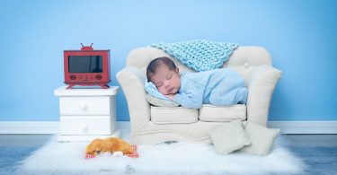 Imagem de um quarto infantil decorado com uma poltrona brnaca, um tapete de pelo, um criado de duas gavetas com uma TV de pequena polegada sobre ele. No sofá temos um recém nascido dormindo bem gostoso.