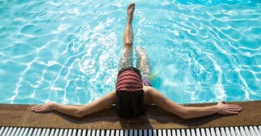 Imagem de uma piscina com uma água azul e limpa. Dentro dela uma mulher encostada na beirada relaxando as suas pernas.