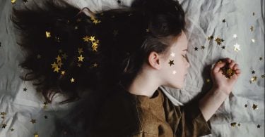 Imagem de uma linda garotinha usando um vestido marrom. Ela está dormindo em uma cama forrada com um lençol branco e sobre ela várias estrelas douradas.