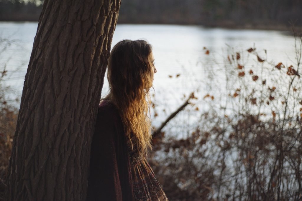 Imagem de fundo contendo um lindo lago. Em destaque uma moça de cabelos longos encostada em uma árvore, ela está pensativa e olhando para o lago.
