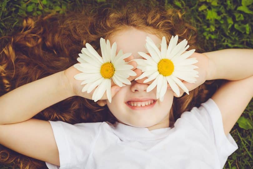 Menina branca segurando flores em seus olhos enquanto sorri.