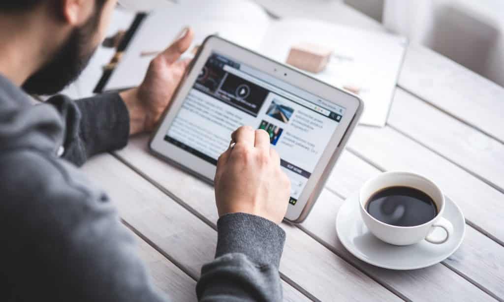 Homem lê site de notícias em tablet. Ele está sentado à mesa e, sobre ela, há uma xícara de café. 