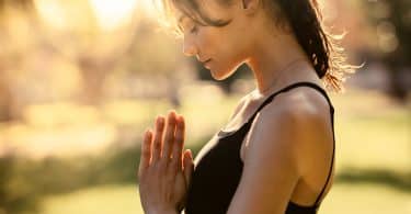 Mulher branca com as mãos juntas e cabeça baixa meditando.