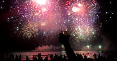 Fogos de artifício luminosos em meio à escuridão. Embaixo deles e em primeiro plano, pessoas de braços erguidos e comemorando.