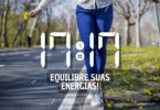 Hora igual 17:17 e texto "Equilibre suas energias!" aplicado por cima de foto das pernas de uma mulher que veste uma calça azul. Ela se equilibra na guia de uma calçada que está coberta de gramas e de flores amarelas. Ela segura pequenas flores amarelas em uma mão. Ao fundo, há uma rua e árvores.