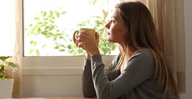 Mulher tomando café e olhando por uma janela.