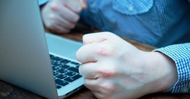 Mão masculina batendo em uma mesa ao lado de um notebook.