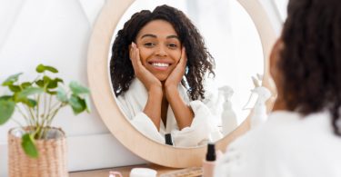 Mulher negra de cabelo cacheado sentada em uma penteadeira e se olhando no espelho, sorrindo