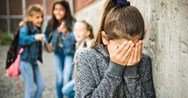 Trio de garotas rindo e apontando para uma menina sozinha encostada na parede com as mãos no rosto chorando