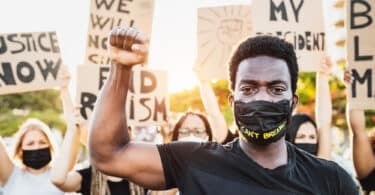 Homem negro parado fazendo o símbolo da resistência com a mão enquanto há pessoas atrás com cartazes em prol do Black Lives Matter