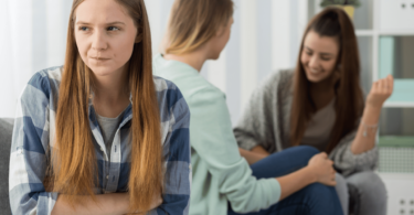 Imagem de três garotas. Duas estão conversando e a outra está irritada, ciumenta e com inveja das duas que batem papo e se distraem.