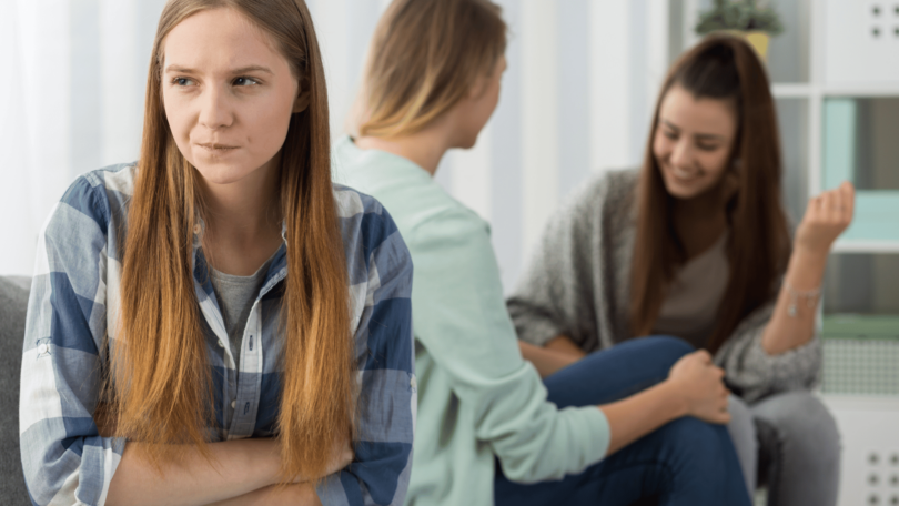 Imagem de três garotas. Duas estão conversando e a outra está irritada, ciumenta e com inveja das duas que batem papo e se distraem.