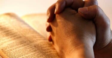 Imagem de uma bíblia aberta e sobre ela as mãos de um homem fazendo uma oração.