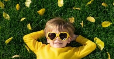 Imagem de uma criança loira, usando uma camiseta de manga longa e um óculos em formato de coração na cor amarela. Ela está deitada em um gramado coberto com folhas amarelas caídas de uma árvore.