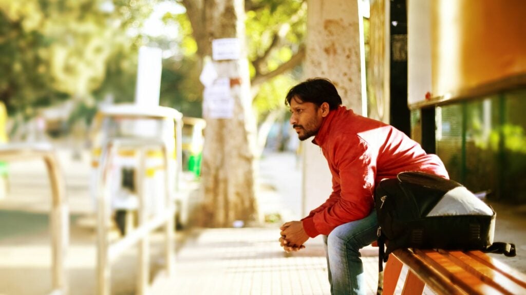 Imagem de um homem usando uma jaqueta na cor vermelha e calça jeans, sentado em um ponto de ônibus.
