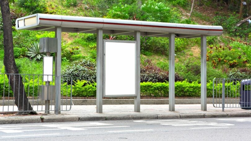 Imagem de um ponto de ônibus vazio. Ao fundo dele, imagens de um jardim.