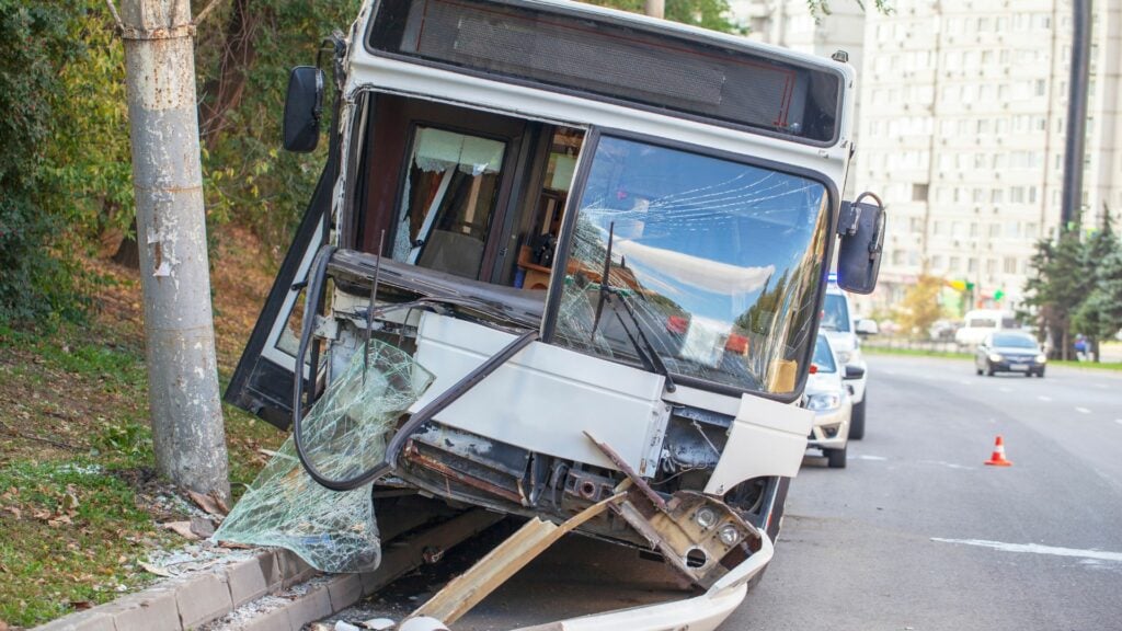 Imagem de um acidente com ônibus urbano de passageiros. O ônibus bateu em um poste em uma rua com pouco movimento.
