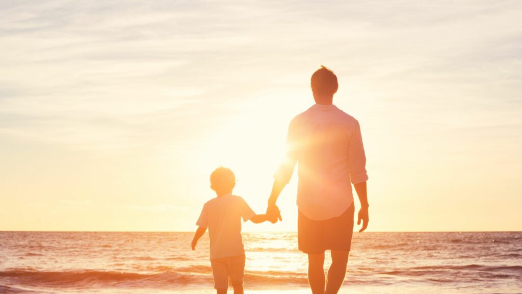 Imagem ao fundo de um mar com um pôr do sol e em destaque a figura do pai e do filho caminhando de mãos dadas na areia da praia.
