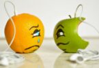 Imagem de uma laranha e uma maçã verde. Uma está bonita e a outra já está mordida. Ambas usam um fone de ouvido e estão representando o sentimento de inveja.